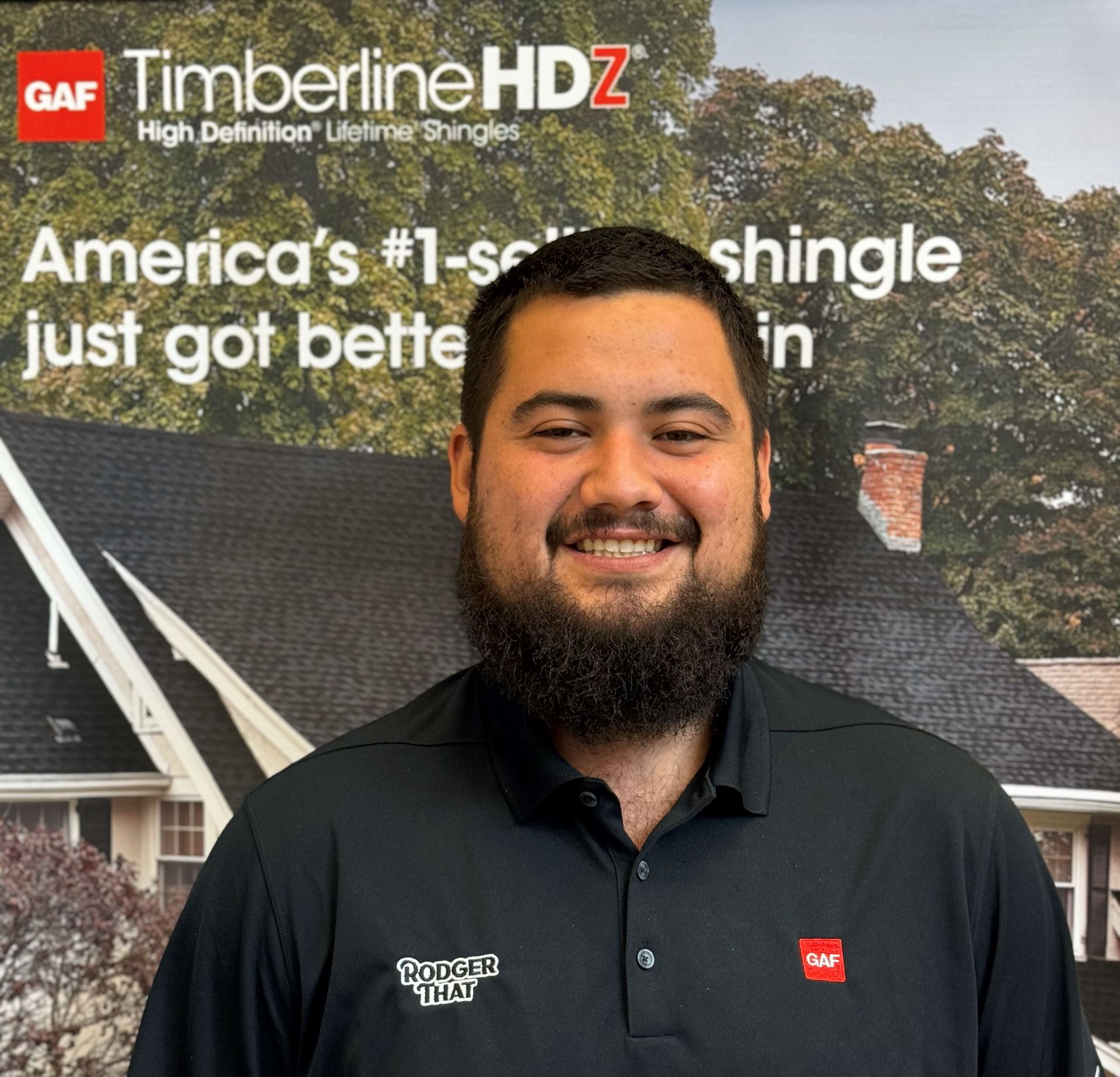 Smiling man in black polo standing before a GAF Timberline HDZ roofing display backdrop