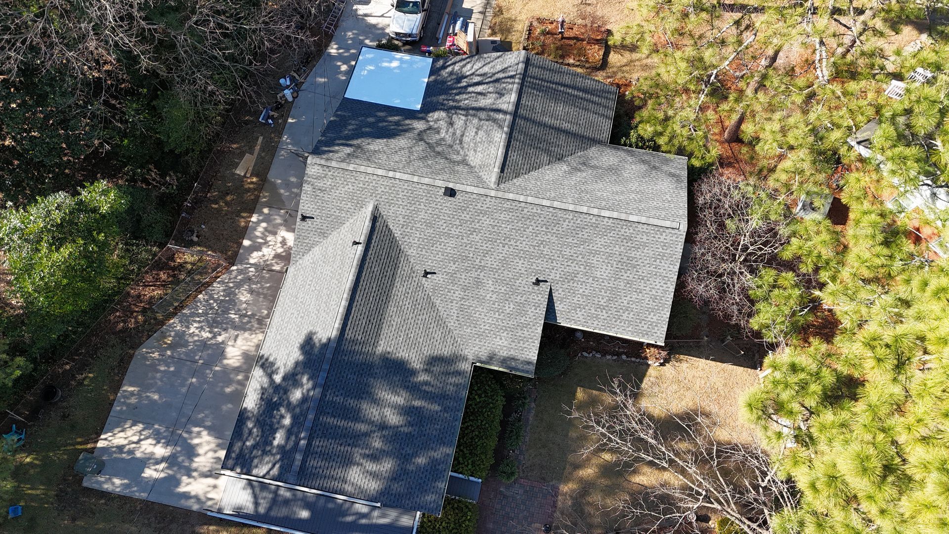 Aerial view of a house roof with dark shingles, skylights, and trees surrounding it.