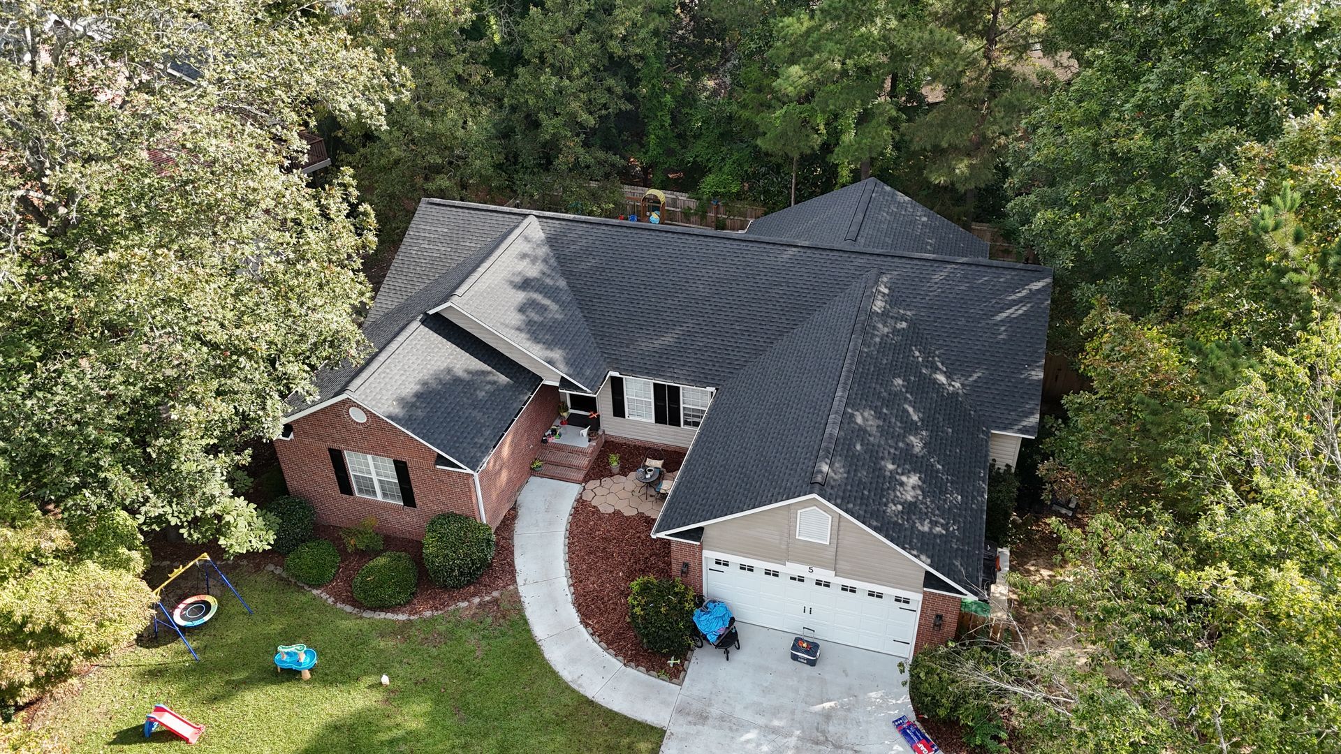 Aerial view of a brick house with a dark roof, curved walkway, and green trees surrounding it