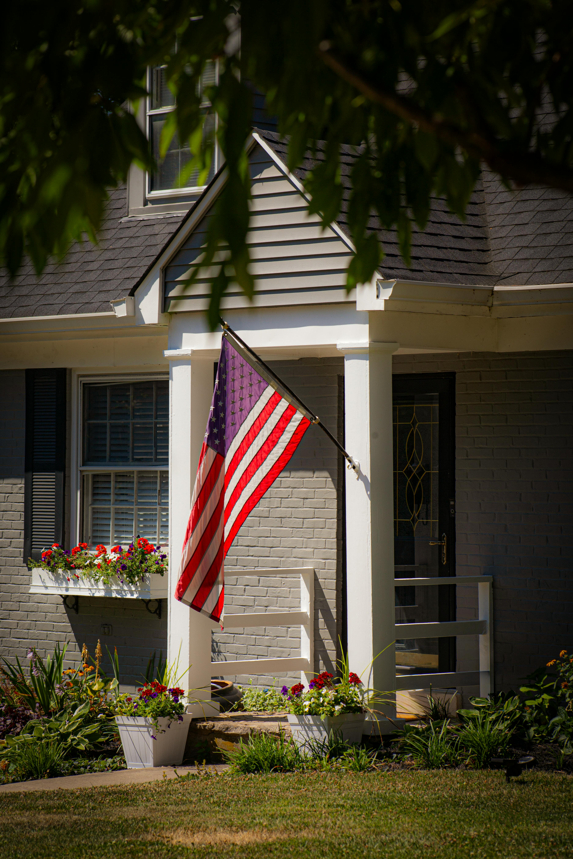 American flag hanging from a porch on a house, with white siding and flower boxes.