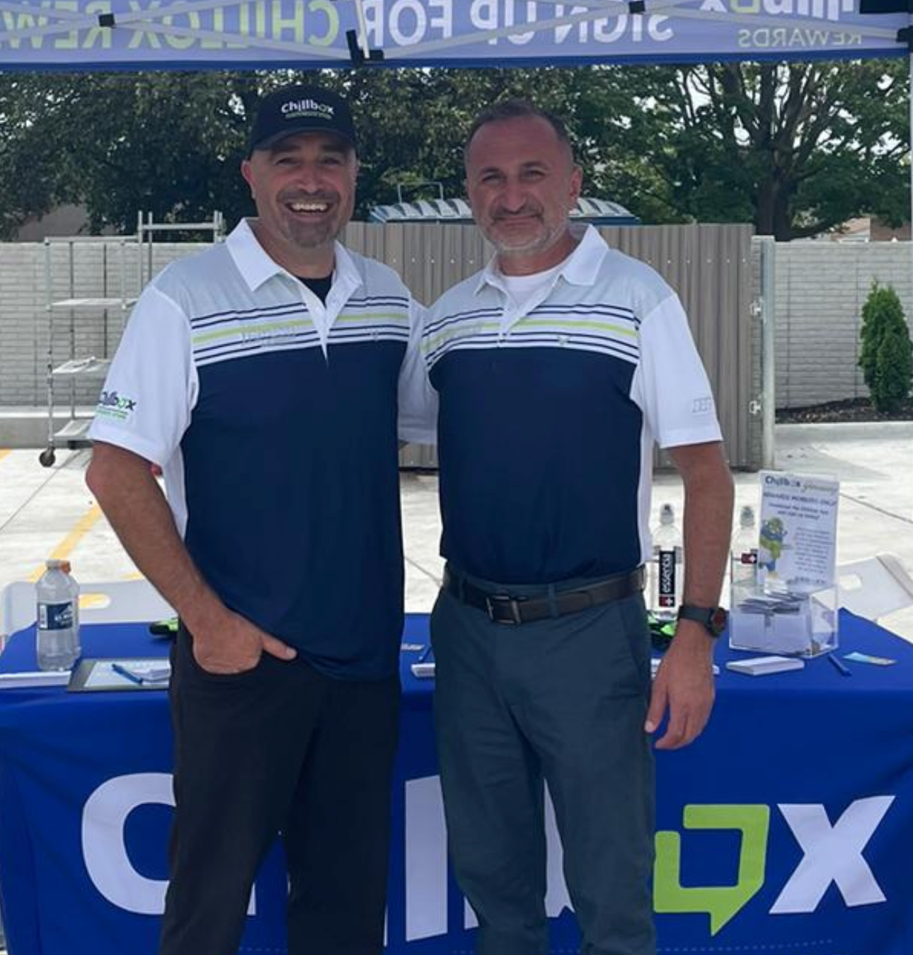 Two men in navy-and-white shirts smiling at a promotional booth under a canopy
