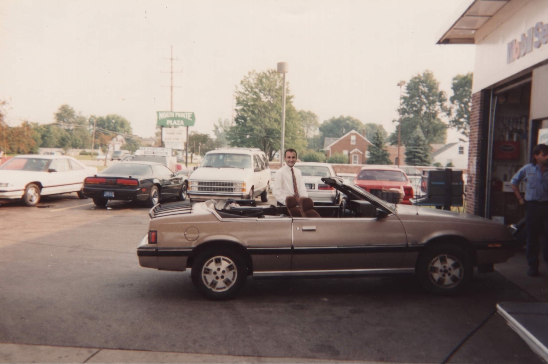 Two men loading a brown sedan at a gas station driveway with parked cars in the background