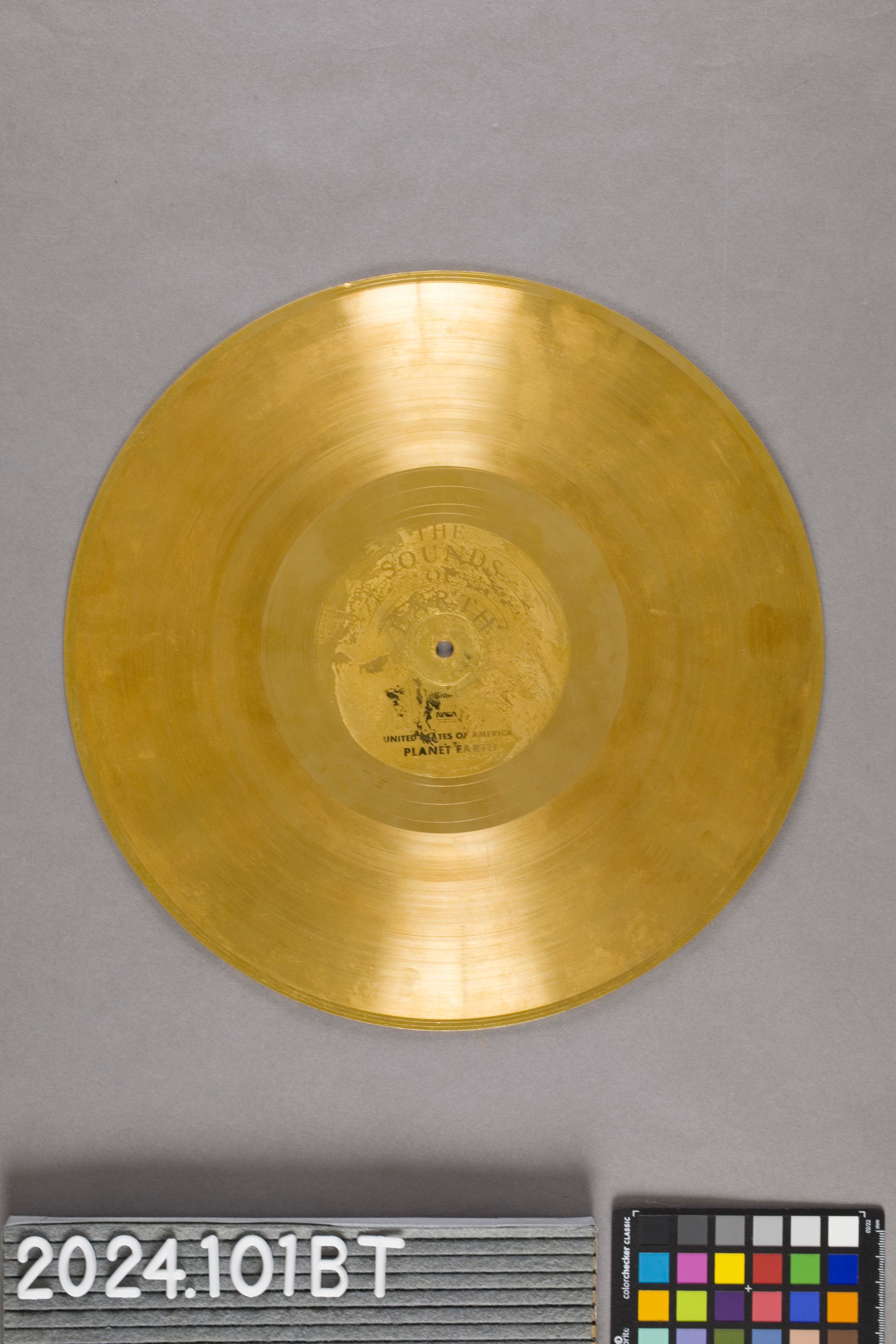 A circular, metallic gold vinyl record centered on a gray background, with a small color calibration chart in the corner.