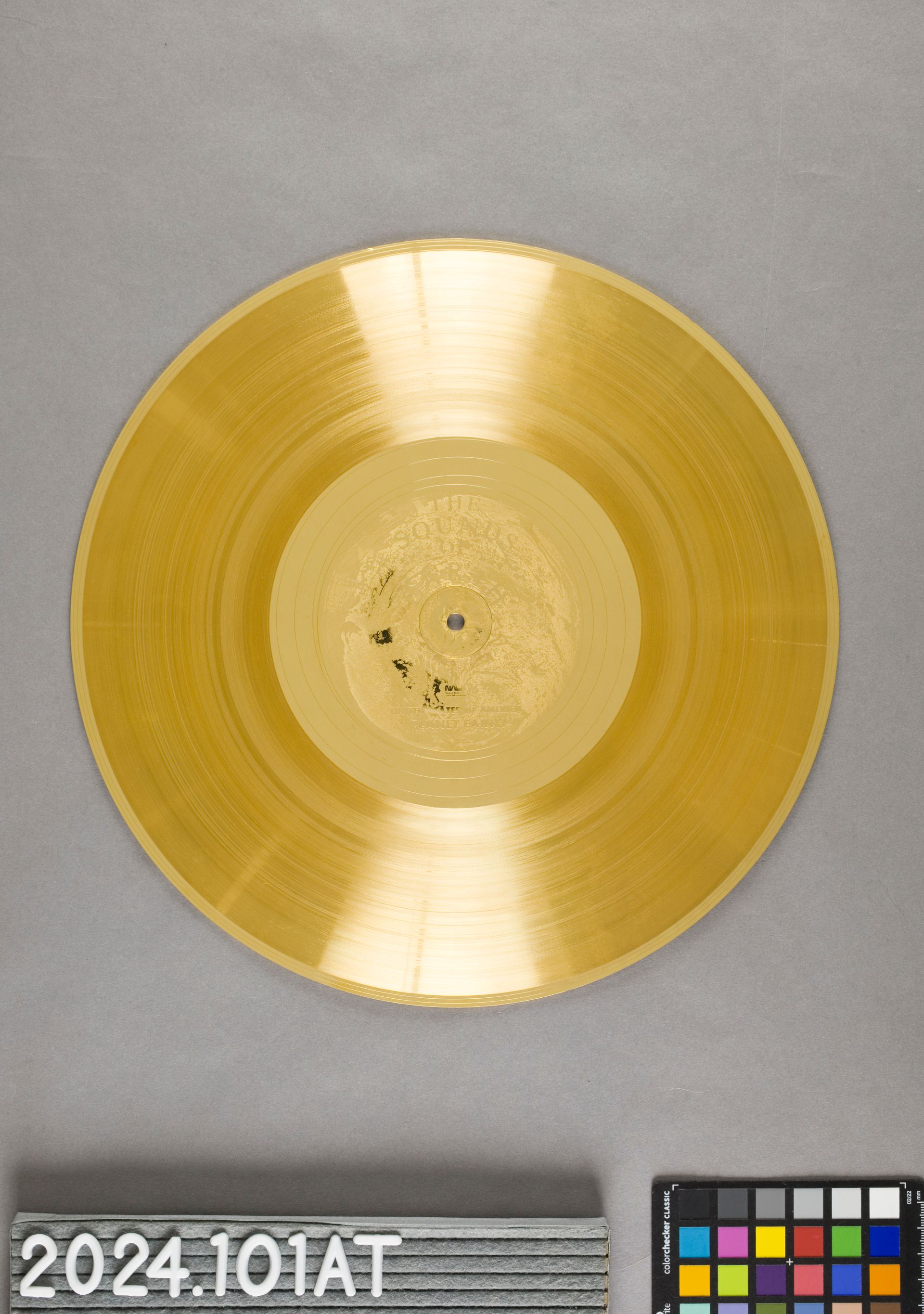 A gold vinyl record on a grey background, next to a reference scale and a color checker card.