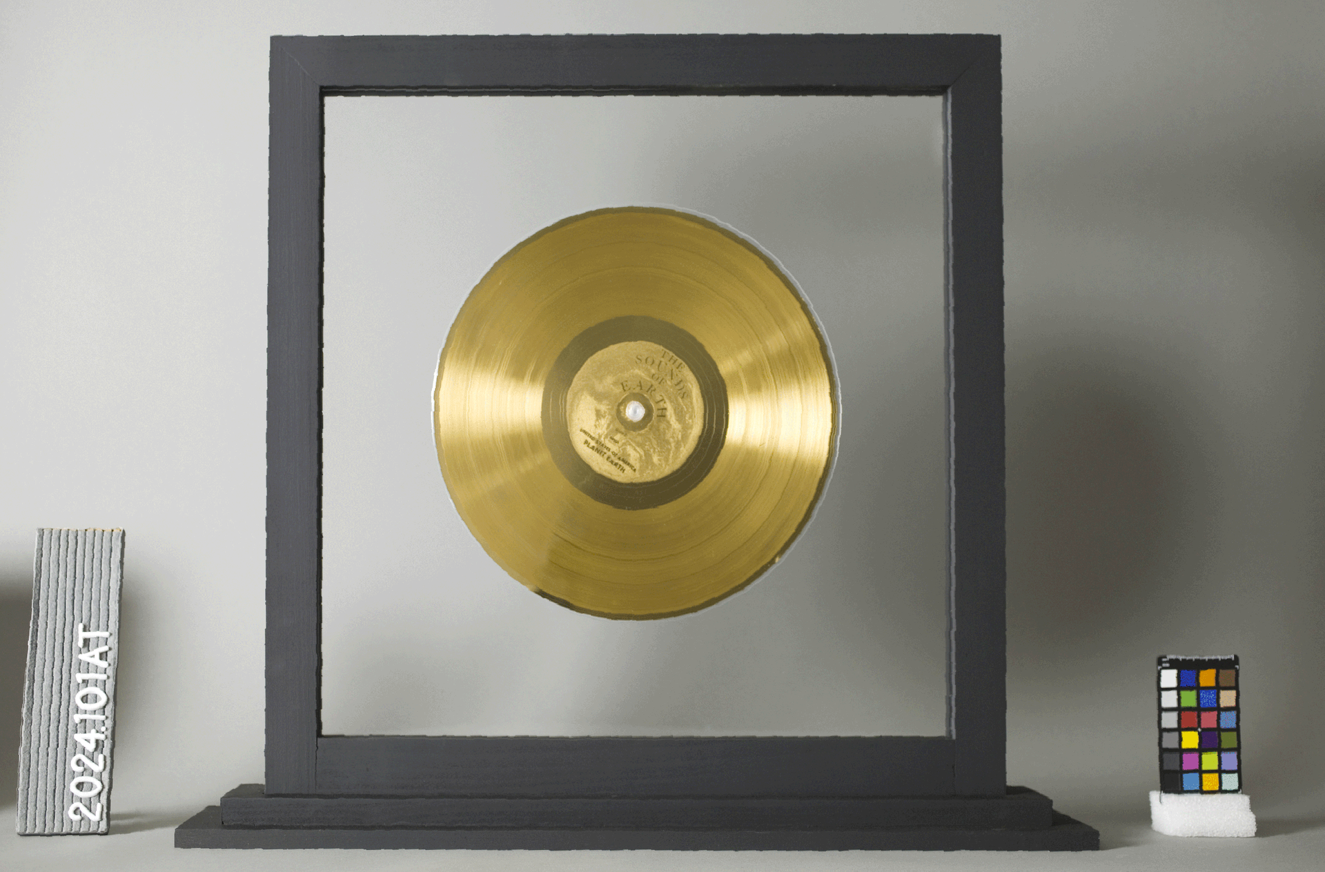 Gold record plaque in a black frame on a shelf, flanked by a small ruler and a color chart.