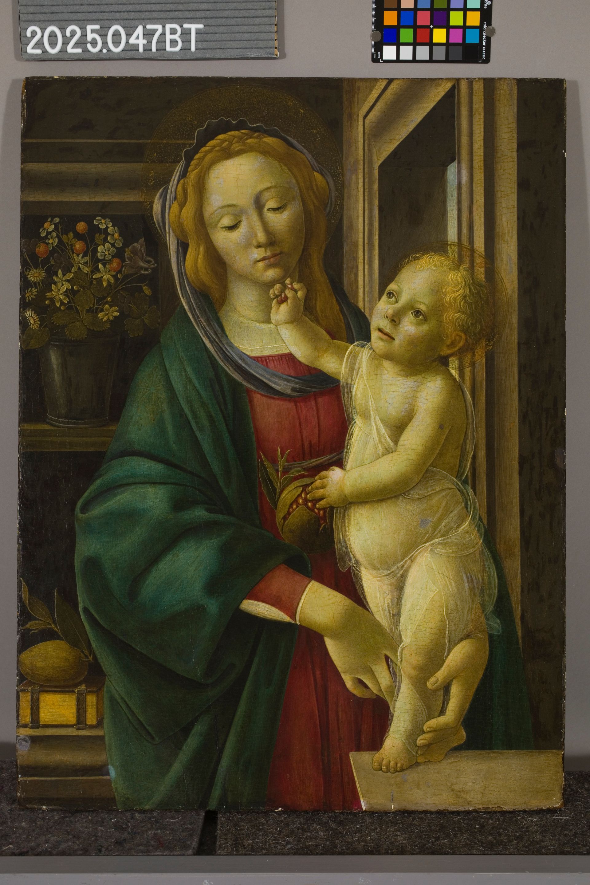 A Renaissance painting of a woman in a red and green robe holding a child who reaches toward her face in an interior.
