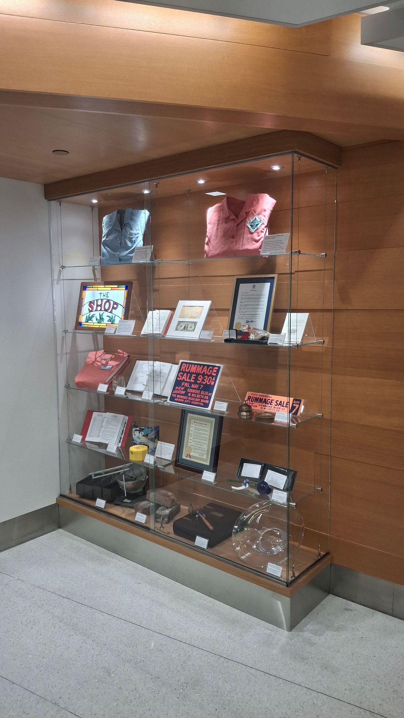 A glass display case with multiple shelves containing clothing, framed items, and various merchandise, set in a lobby.