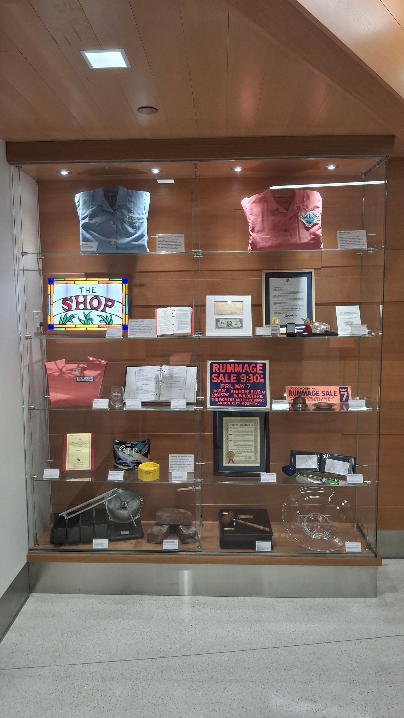 A wooden display cabinet containing memorabilia, framed documents, clothing, and small decorative items on glass shelves.