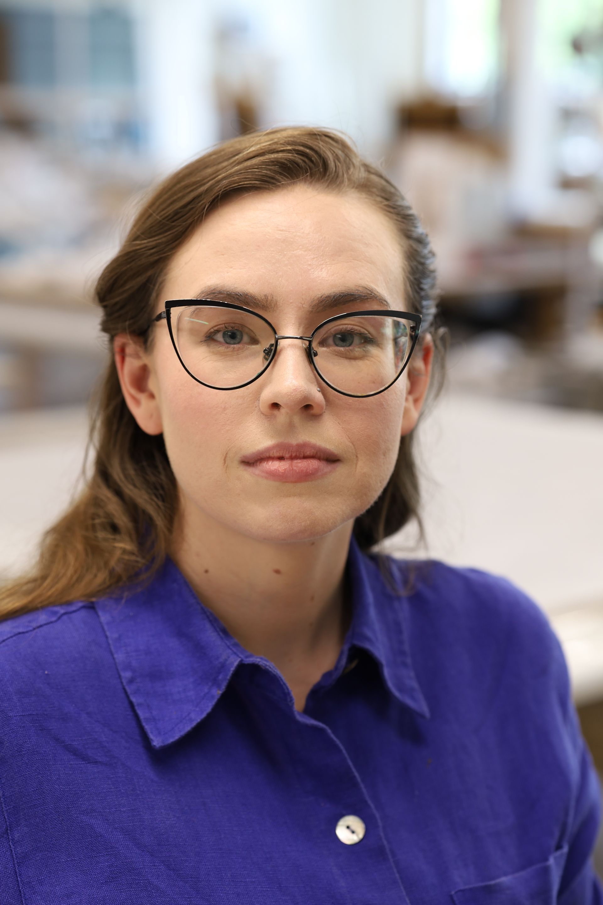 A person with long brown hair wearing cat-eye glasses and a blue button-down shirt, looking forward against a blurred background.