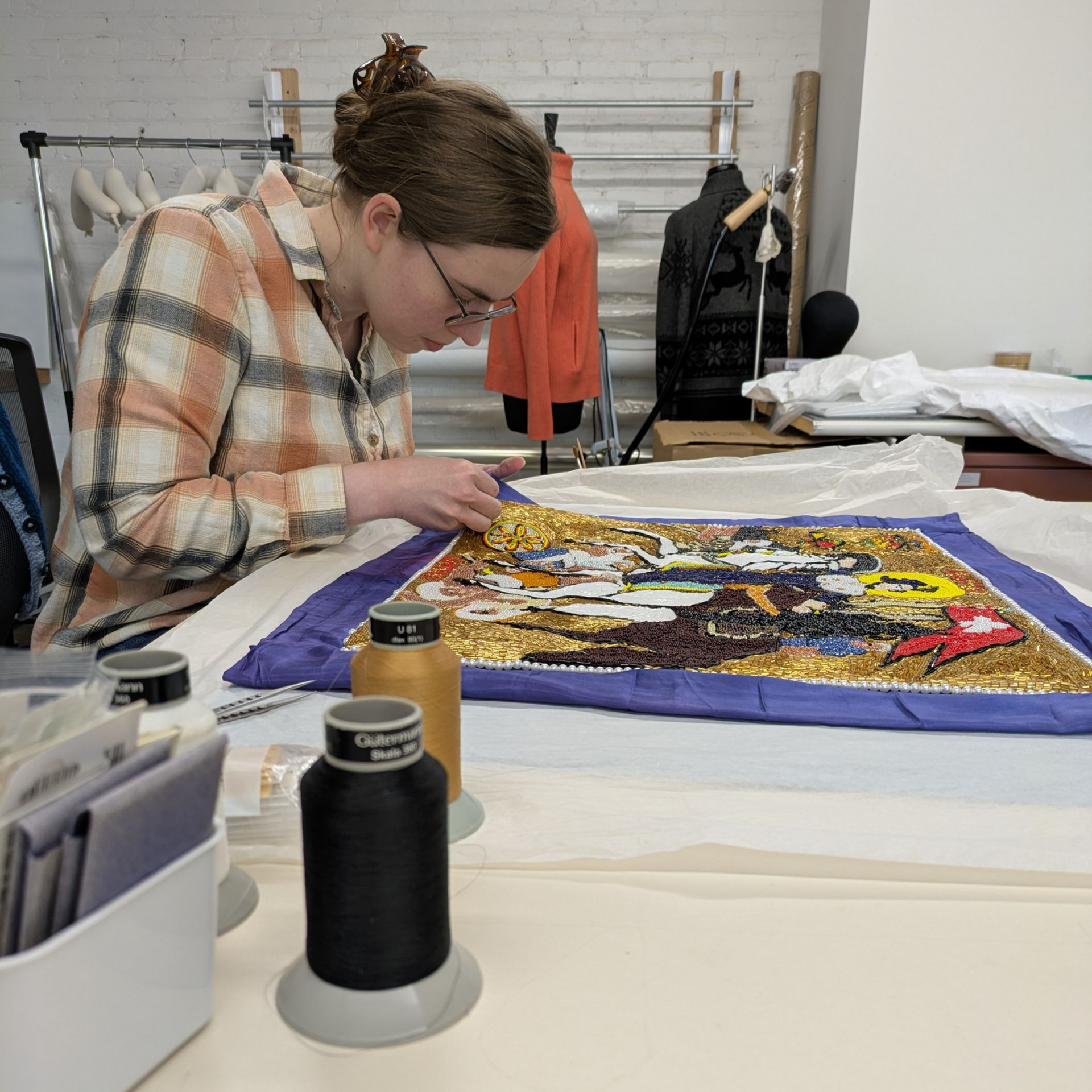 A woman is sitting at a table working on a piece of embroidery.