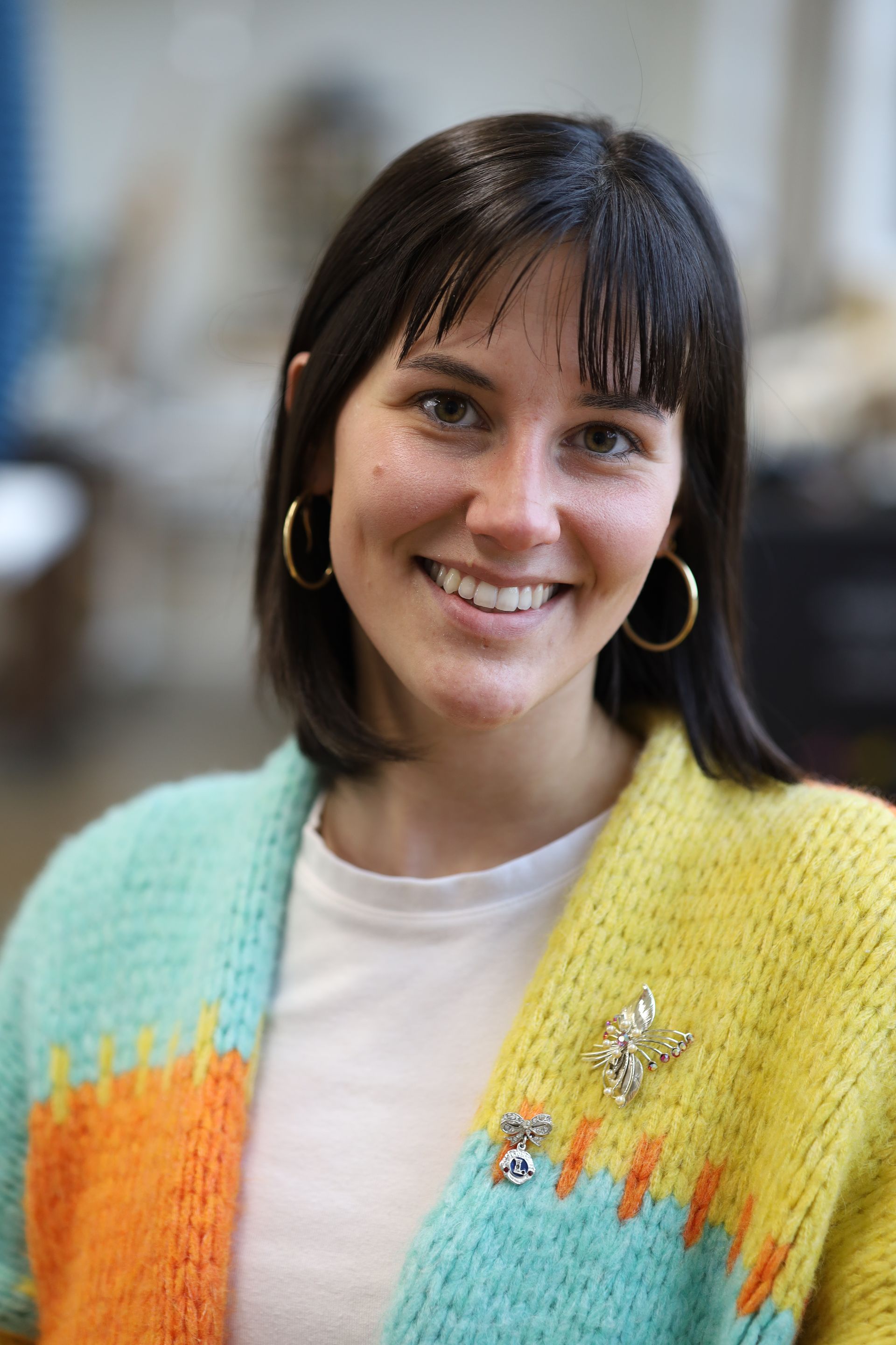 Smiling person with dark hair and gold hoop earrings wearing a multi-colored chunky knit cardigan with two pins.