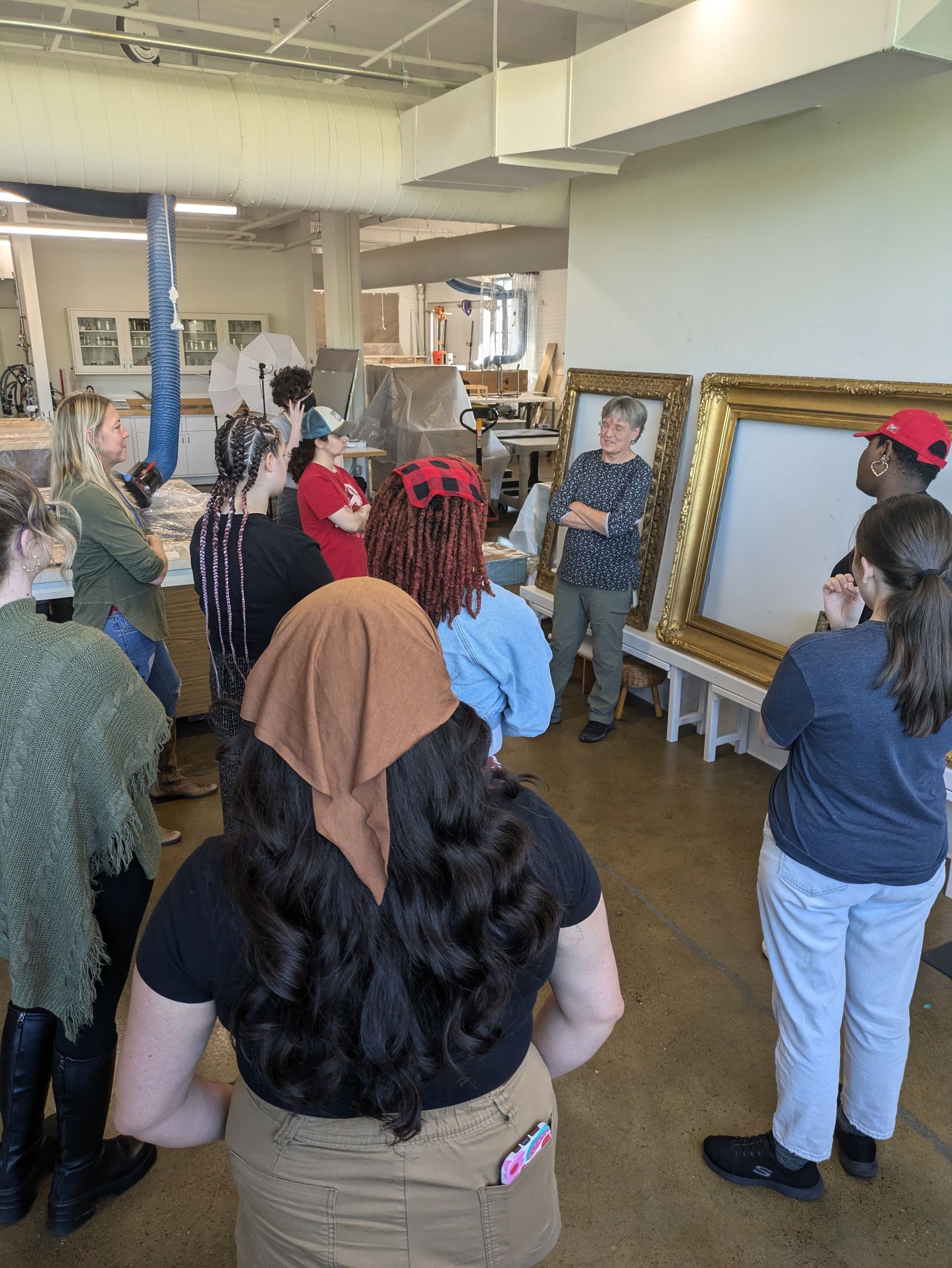 A group stands in an art studio, listening to a person gesturing near two large, ornate gold frames on a table.