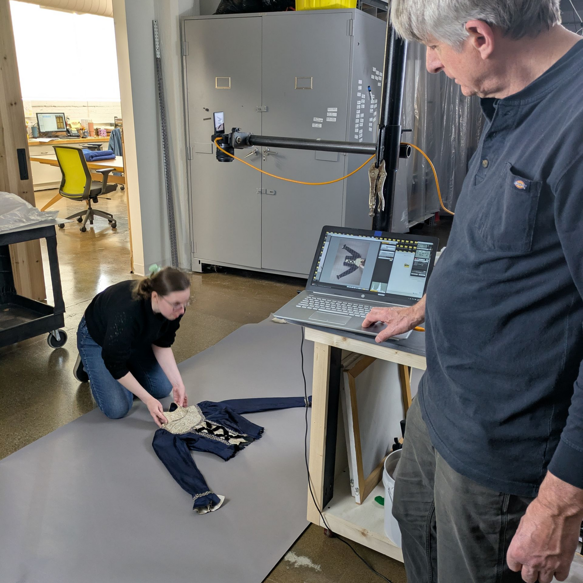 Two people in a workshop examining a mannequin and laptop on a table, with one person crouched on the floor.