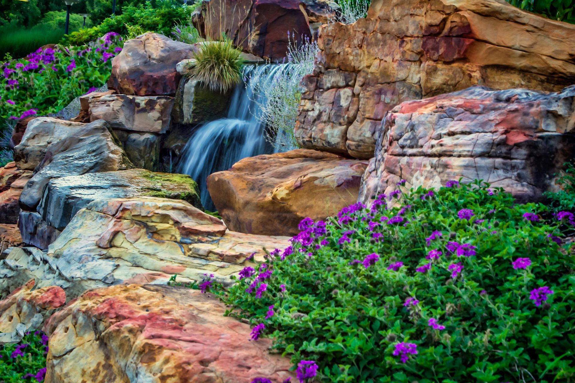 A waterfall surrounded by rocks and purple flowers in a garden.