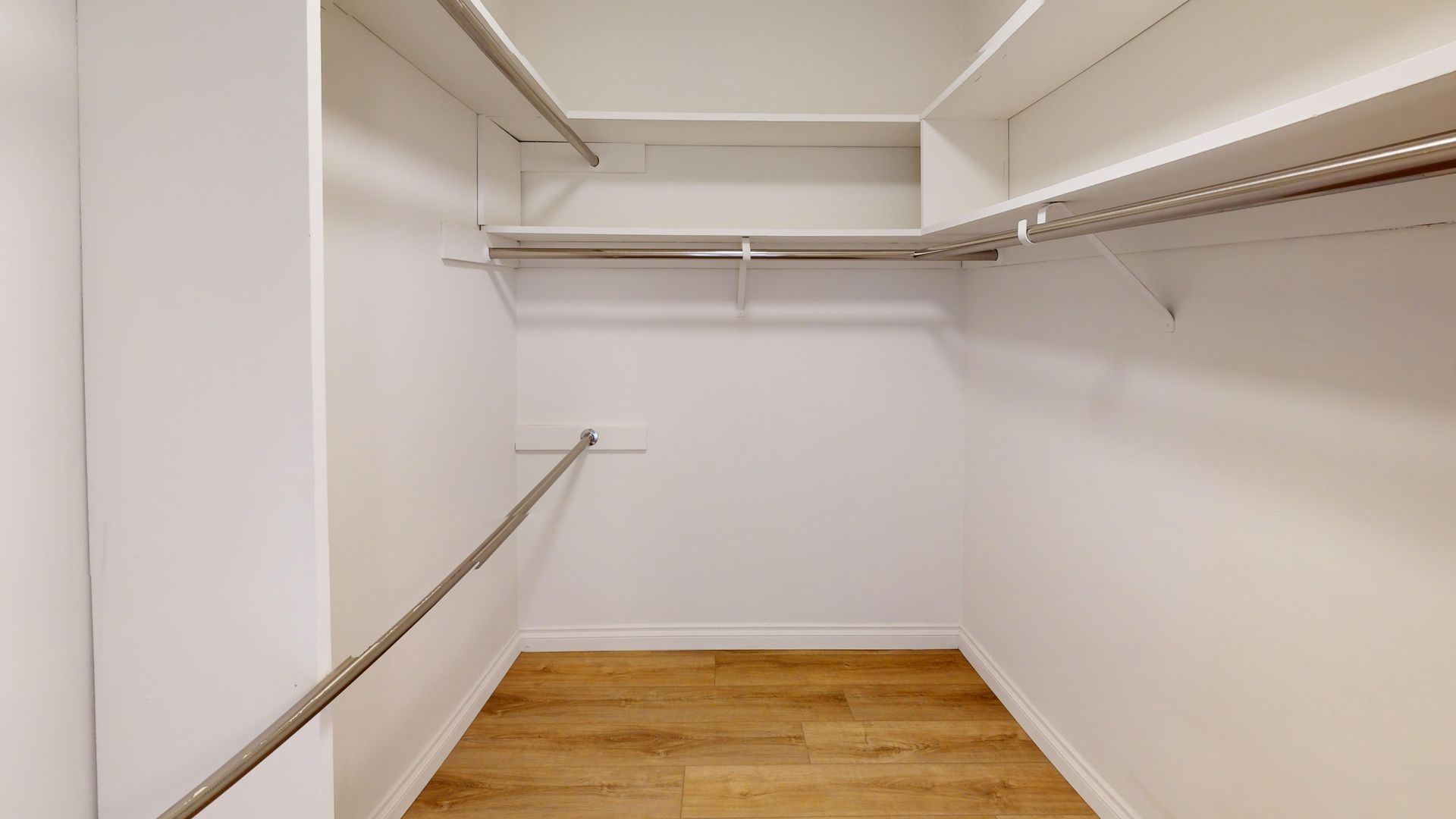 Empty white walk-in closet with shelves and rods, light wood floor.
