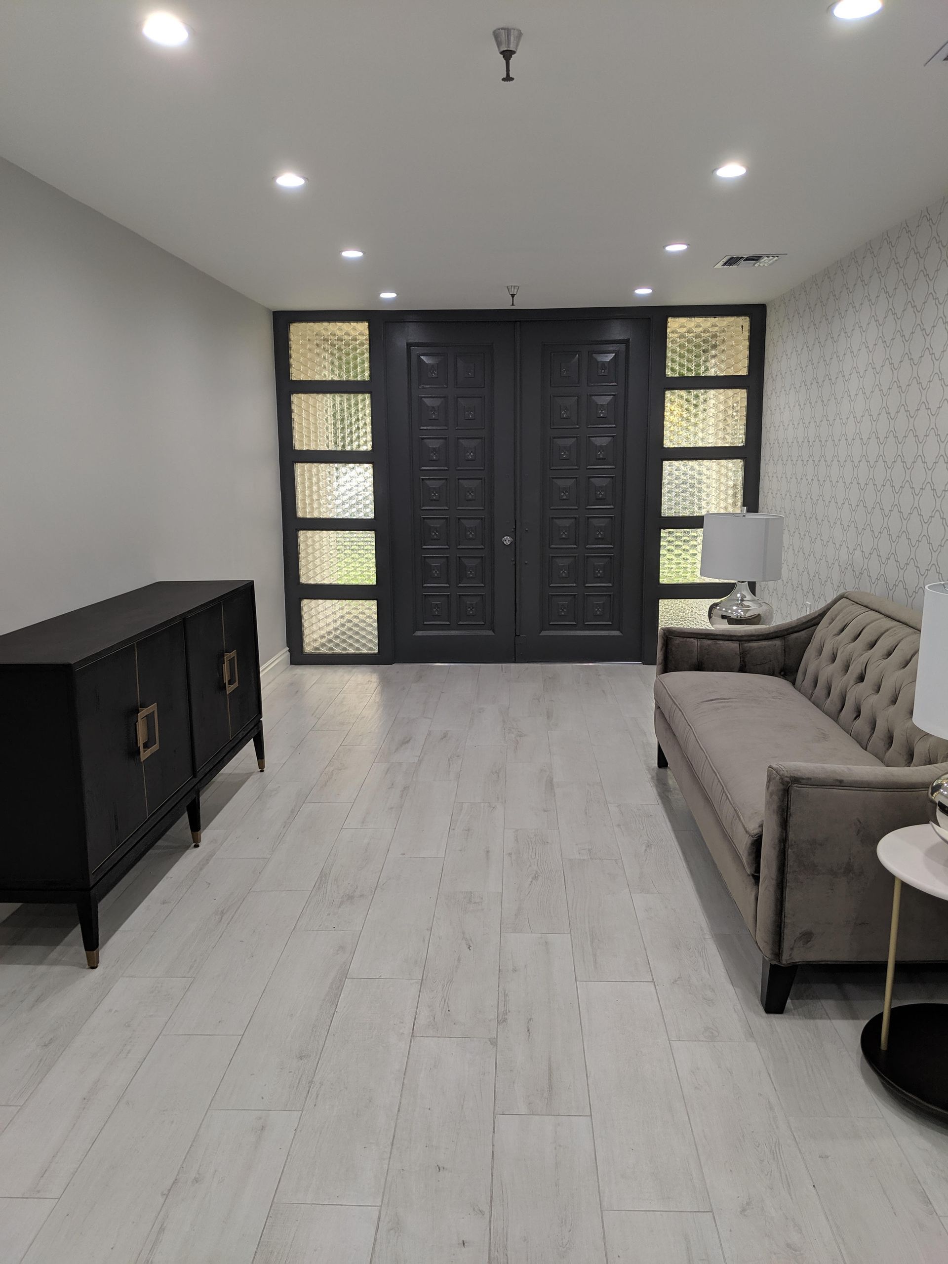 Gray entryway with dark gray double doors, light wood-look floor, black cabinet, gray sofa, and two lamps.