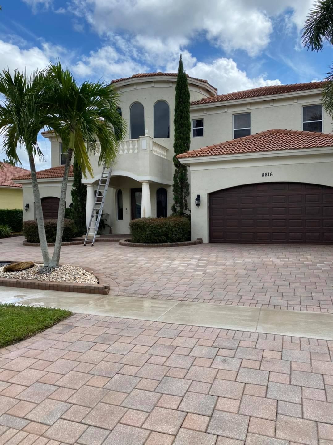 Two-story beige house with red-tile roof and brown garage door on a brick driveway under a blue sky.