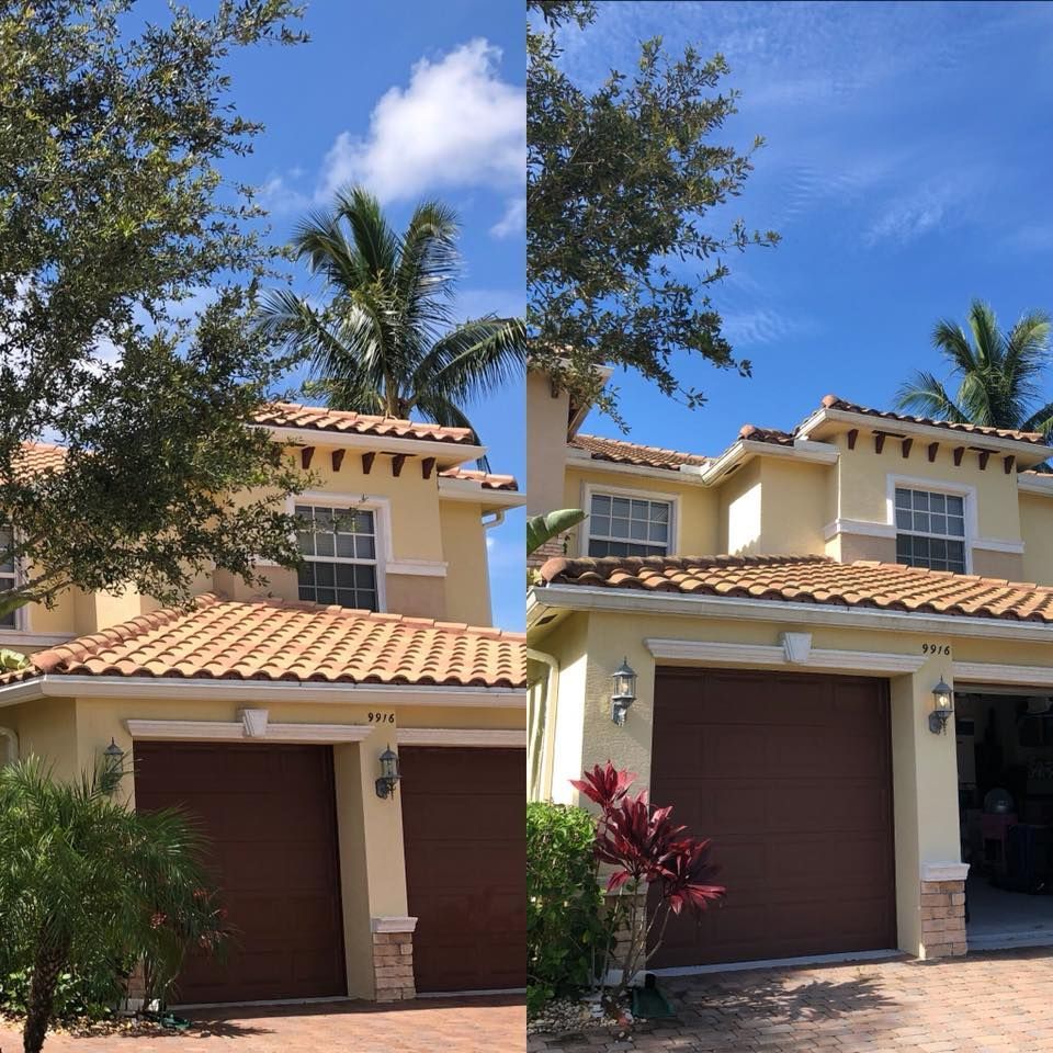 Two-story tan house with brown garage doors under a blue sky, with palm trees and other greenery in the yard.