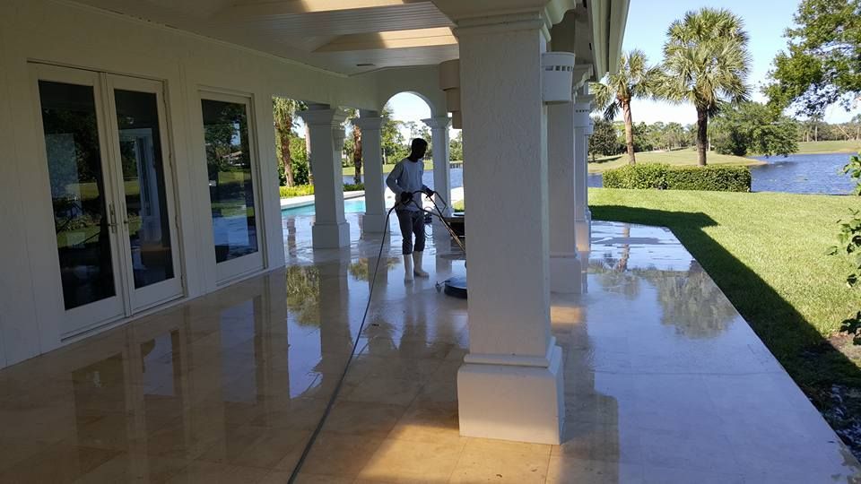 A person cleaning a shiny, light-colored patio with columns and glass doors, overlooking a pool and lake on a sunny day.