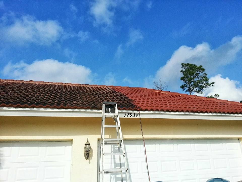 A roof is being cleaned with a pressure washer. The left side is dark with algae, while the right side is bright red.