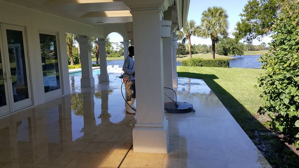 Man pressure washing a polished outdoor patio, overlooking a lake and manicured lawn. White columns support the roof.