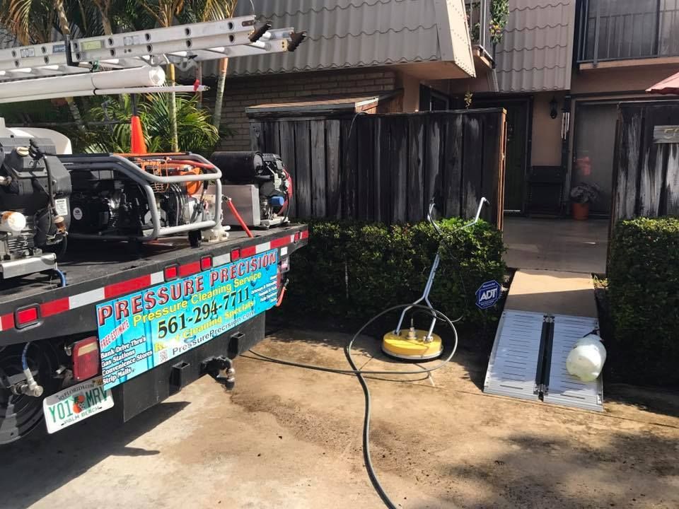 A pressure washing truck cleaning a property. The truck is parked next to a fence and bushes, with equipment deployed.