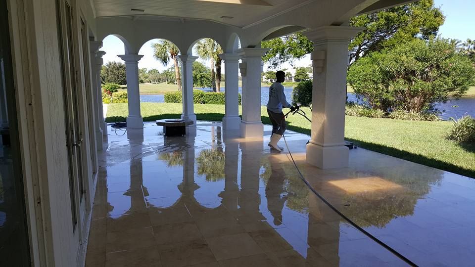 A person pressure washes a gleaming tiled patio, with columns, overlooking a lake and green lawn.