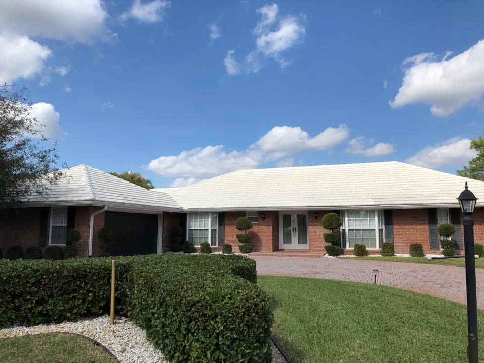Brick ranch house with a white roof, surrounded by green lawn and bushes under a blue sky with clouds.