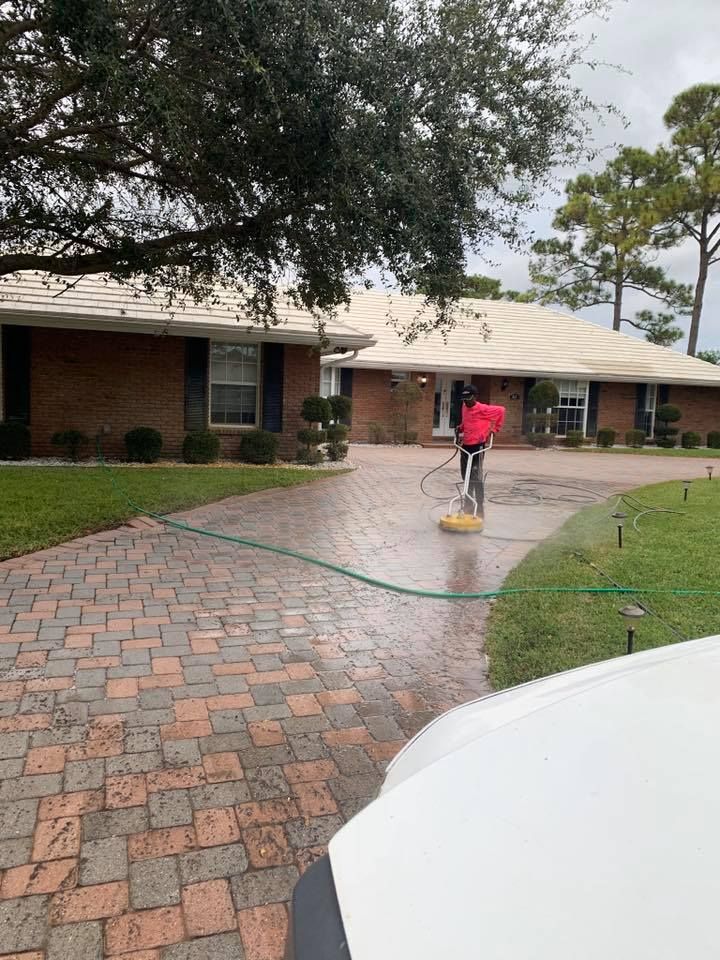 Person pressure washing a brick driveway in front of a brick house with a manicured lawn.