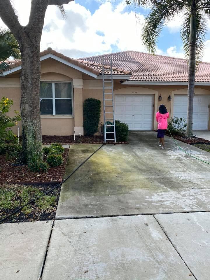 A person in pink stands near a ladder leaning against a tan house with a red tile roof. The driveway is partially cleaned.
