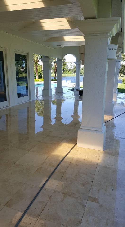 A covered outdoor patio with reflective, light-colored tile flooring. Columns and a pool are visible in the background.
