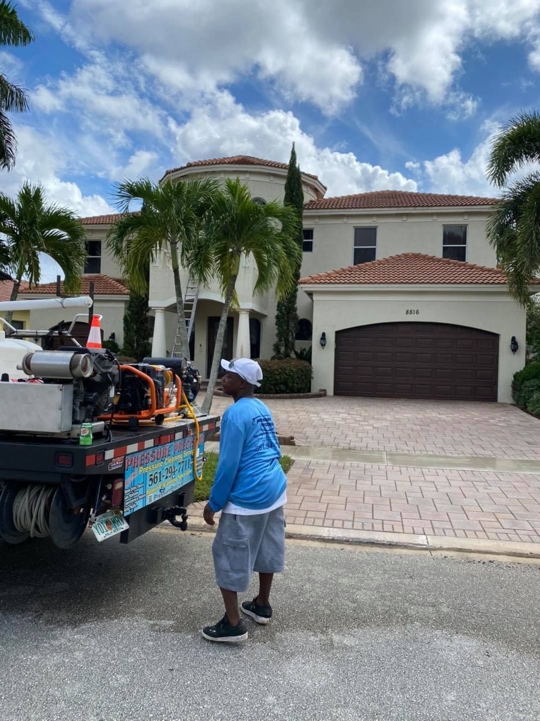 Man in a blue shirt stands near a truck with equipment in front of a large house with a red-tiled roof on a sunny day.