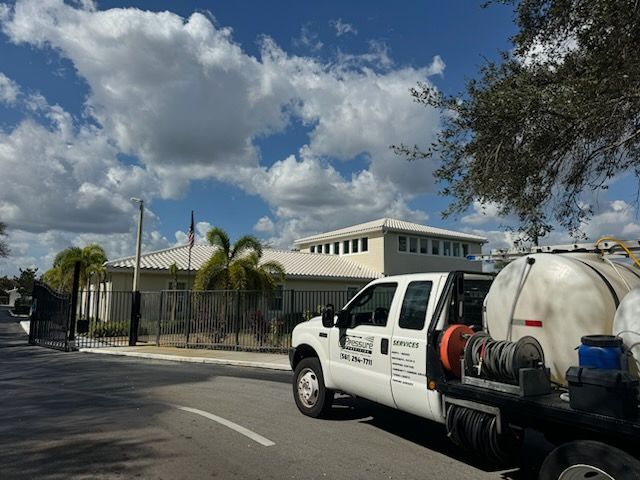 A white truck with a water tank parked on a street in front of a light-colored building under a partly cloudy sky.