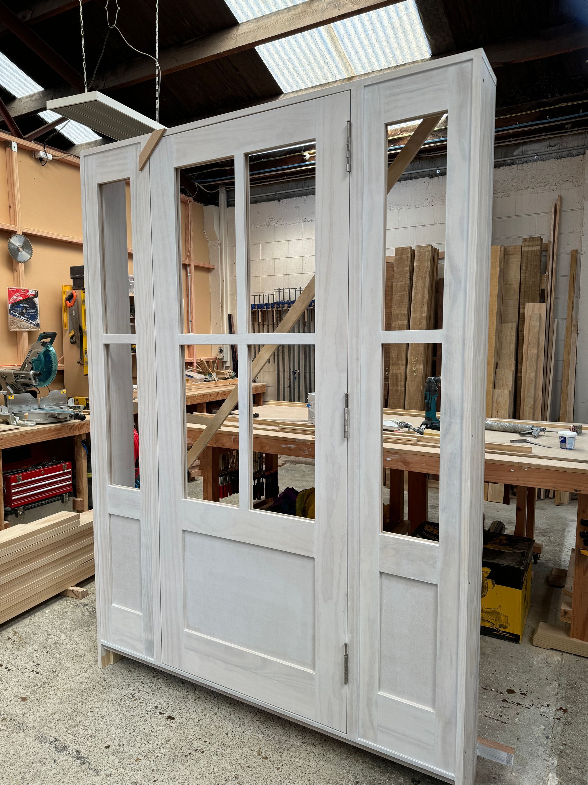 A white-painted, three-panel wooden screen with glass windows and solid base panels, standing in a workshop.
