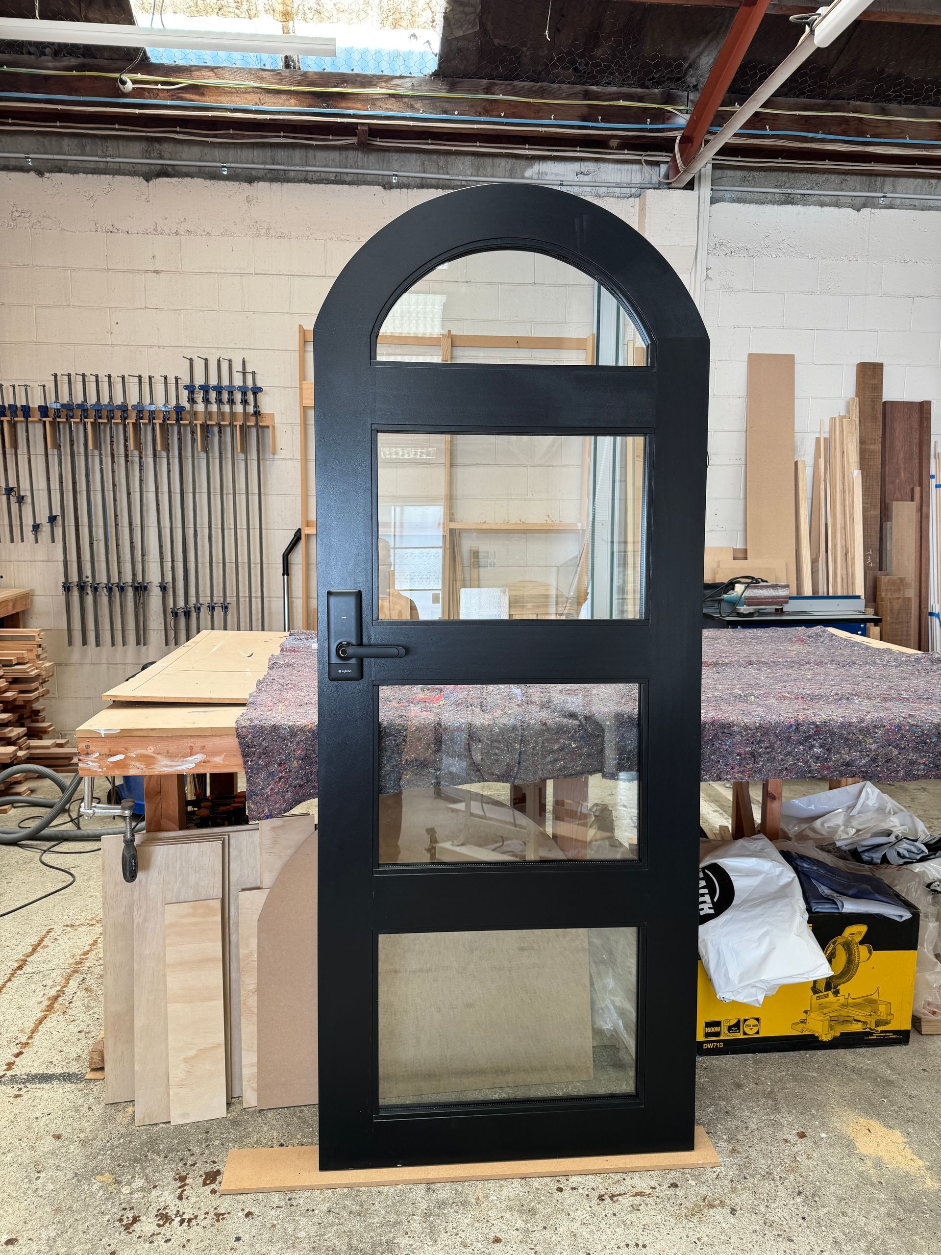 A black arched door with three glass panes stands in a woodworking shop, resting on a wooden board.