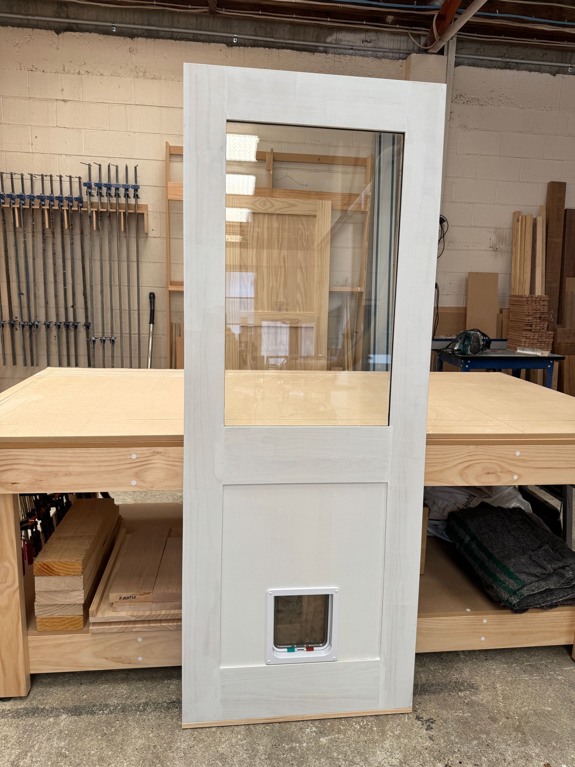 A white door with a glass panel and an integrated pet door standing on a wooden workbench in a workshop.