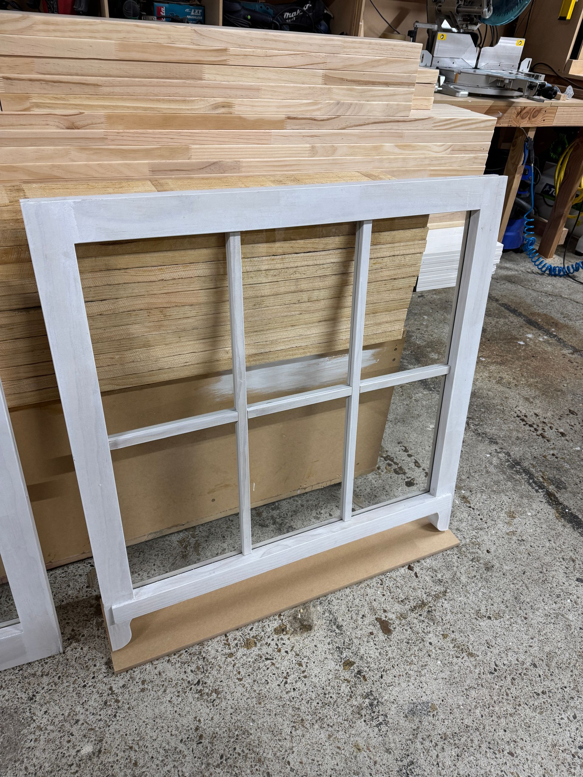 A white, six-pane window frame resting on a wooden board in a woodworking shop.