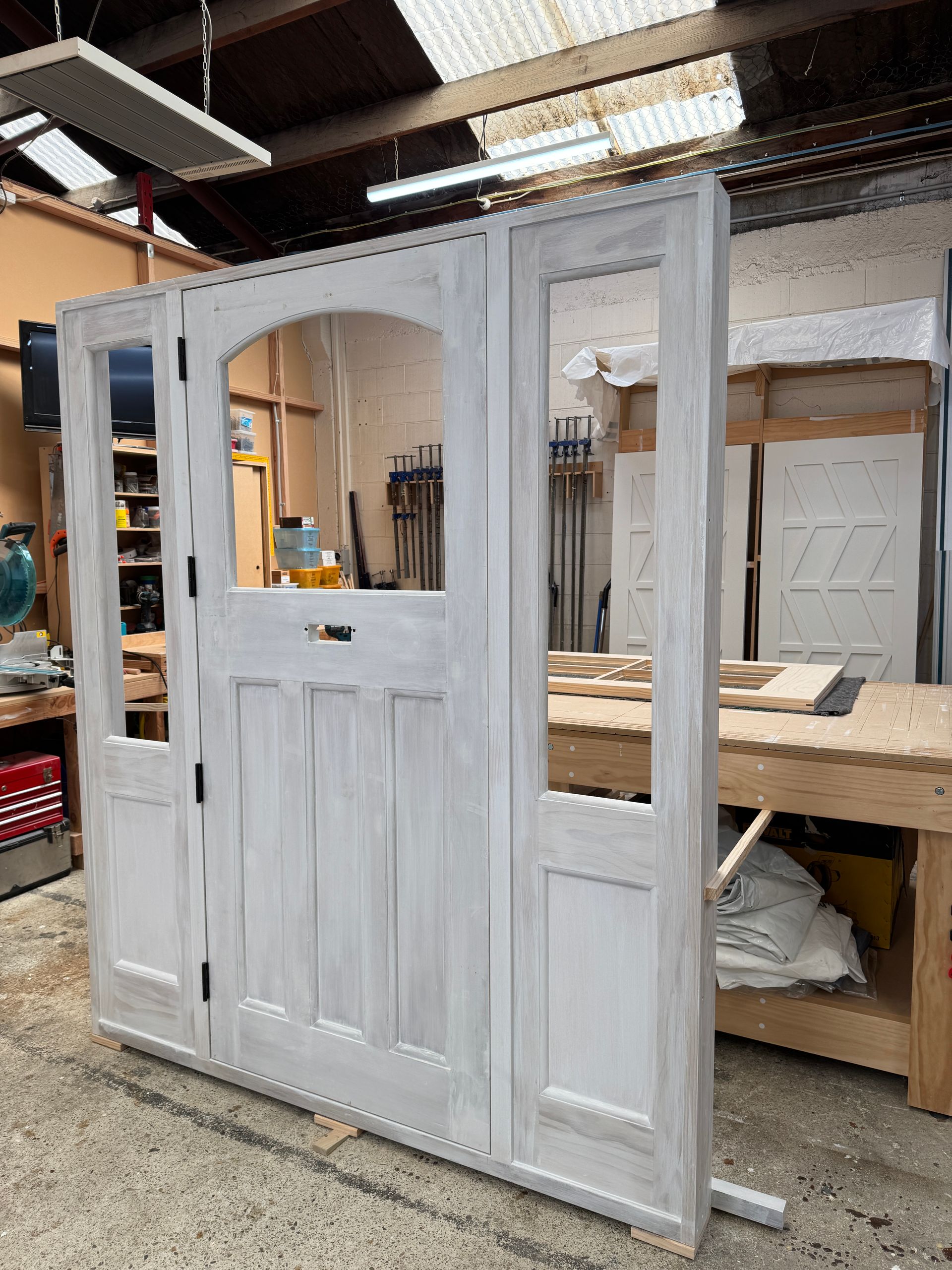 A white, arched wooden front door with side panels and glass windows, standing in a workshop.