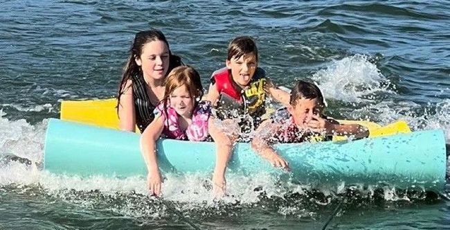 Four children wearing life jackets hold onto a large, light blue floating tube while being pulled through the water.