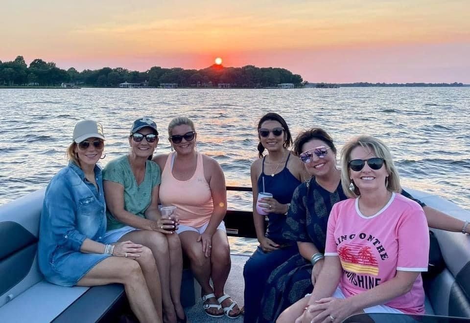 Six smiling people sit on a boat during sunset on a lake, with the sun setting behind a distant tree-lined shore.