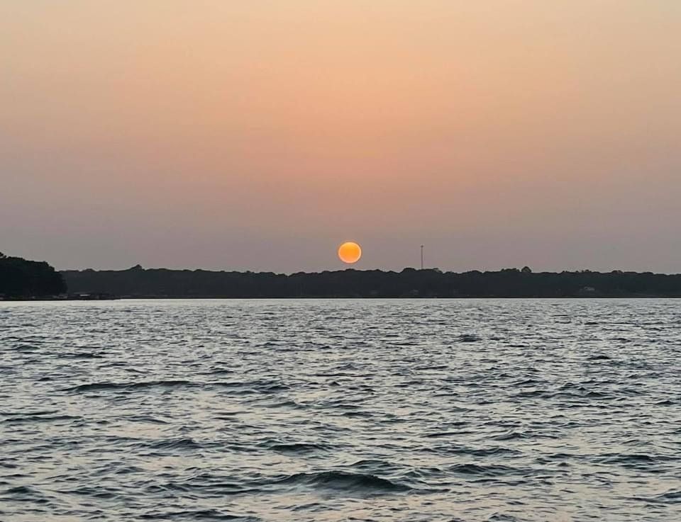 A golden sun sets over a dark, tree-lined horizon above a calm, rippling body of water.