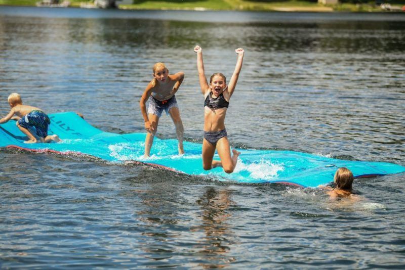 Children play on a bright blue floating mat in a lake, with one person jumping and cheering in the center.
