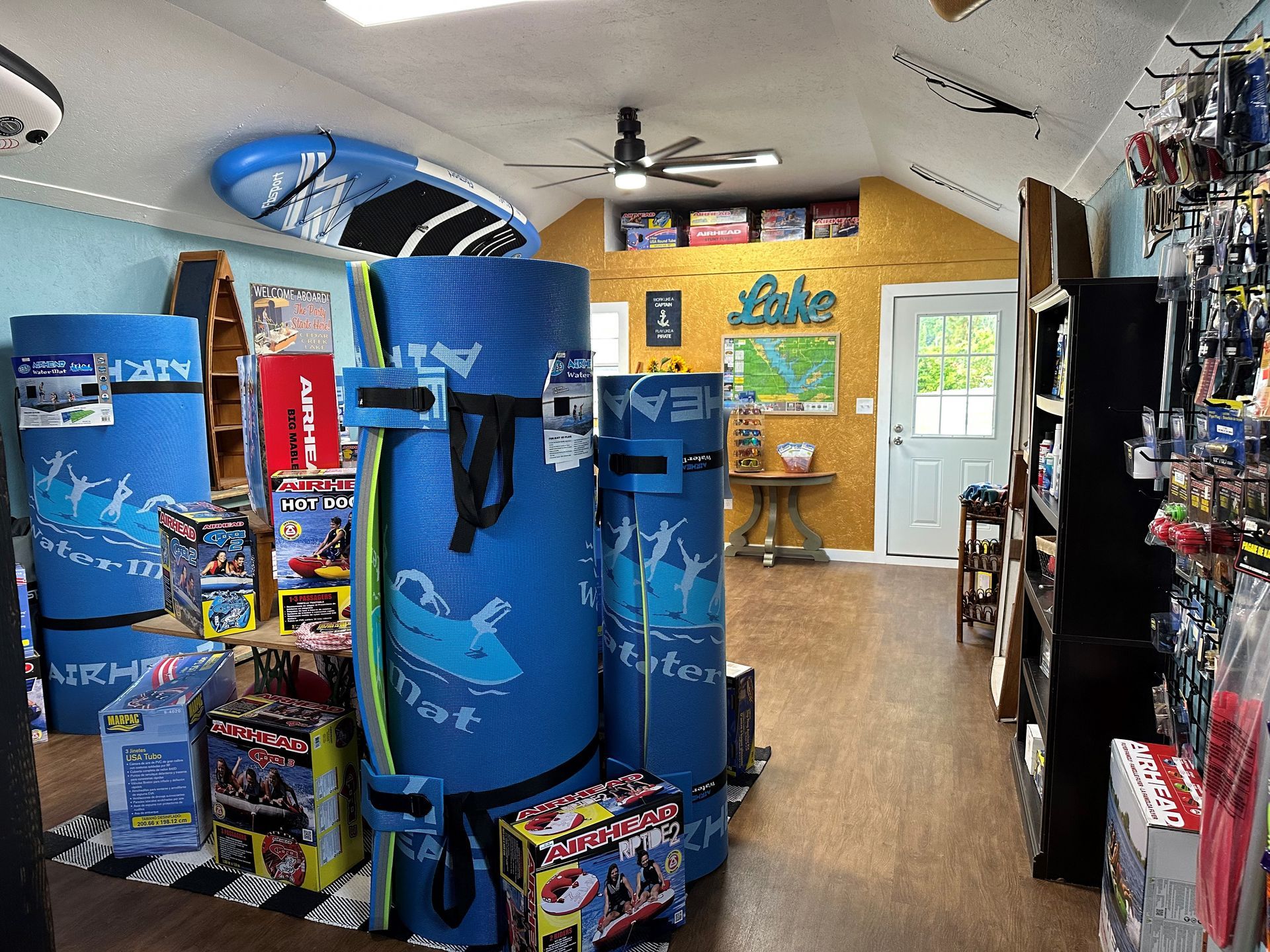 An indoor shop displaying blue inflatable water mats, paddleboards, and retail items, with a door leading to the outdoors.