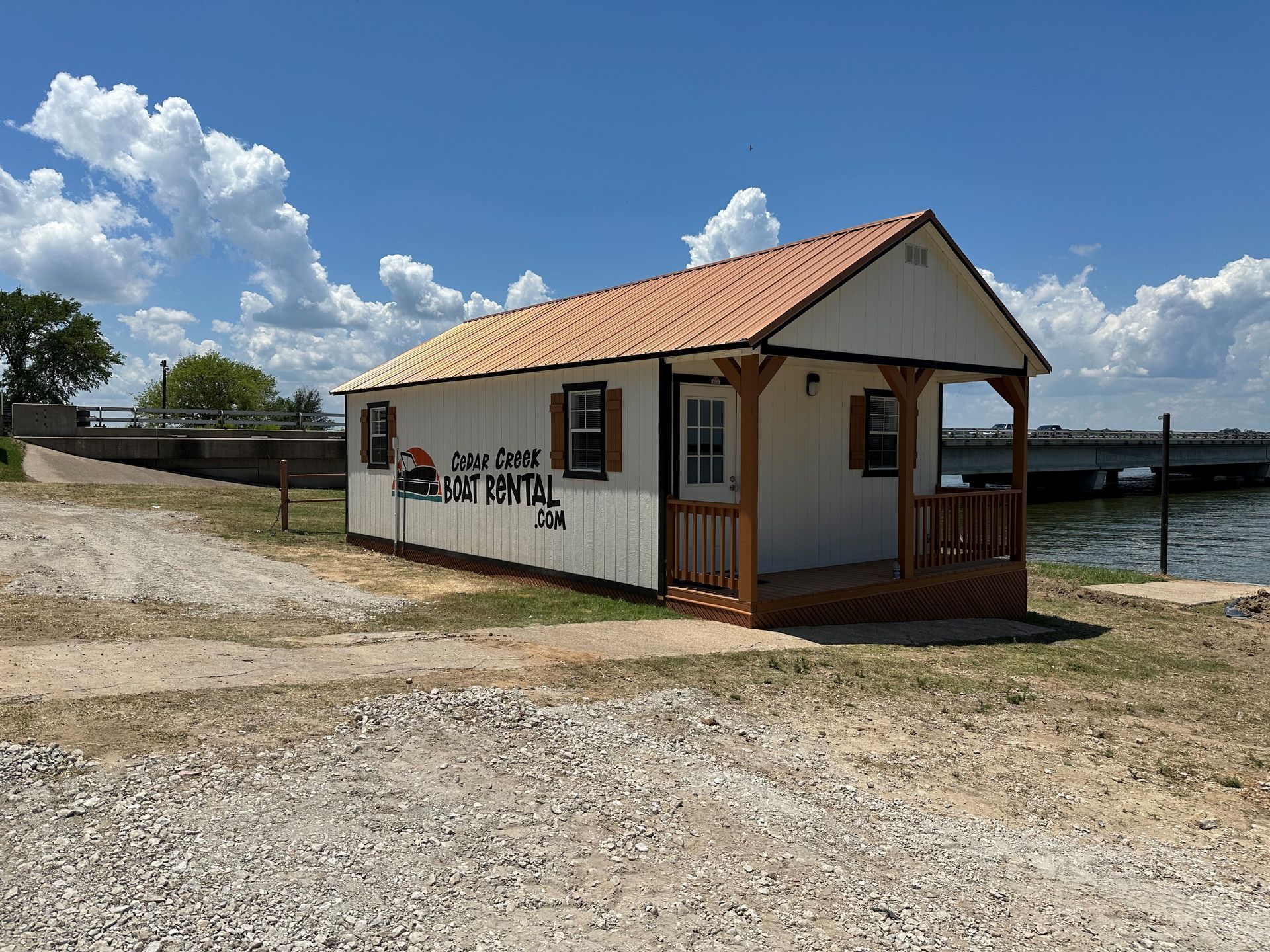 A white boat rental shack with a tan roof and wooden porch stands by a lake near a bridge under a blue sky with clouds.