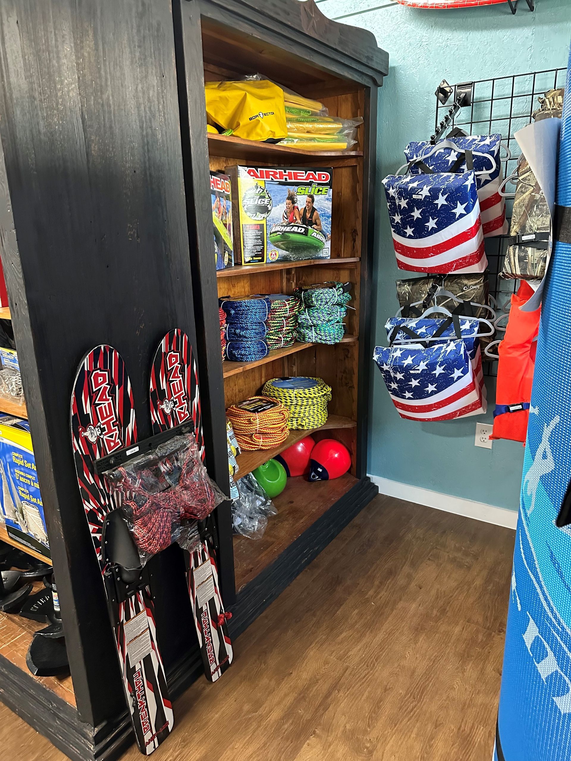 A wooden display shelf in a store holds toys and sports gear, with patterned bags hanging on the wall beside it.