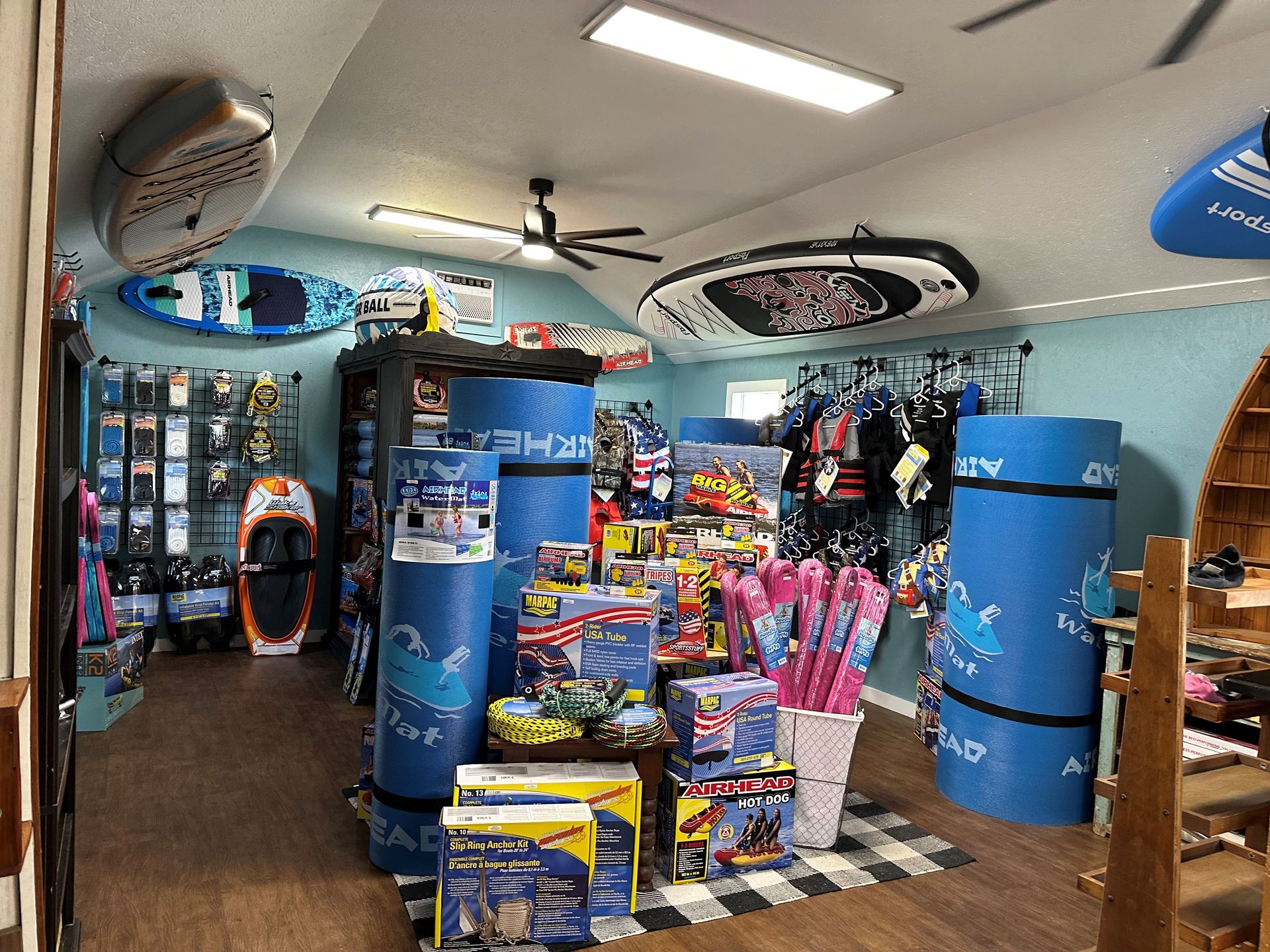 An indoor retail shop filled with water sports equipment, including paddleboards, racks of apparel, and boxes of toys.