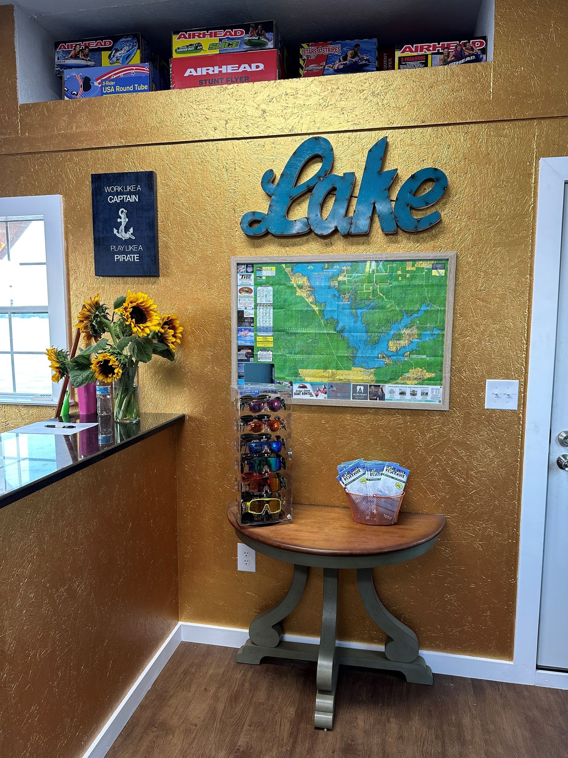 A room features a gold glitter wall with a teal “Lake” sign, a map, a side table with sunglasses, and sunflowers in a vase.