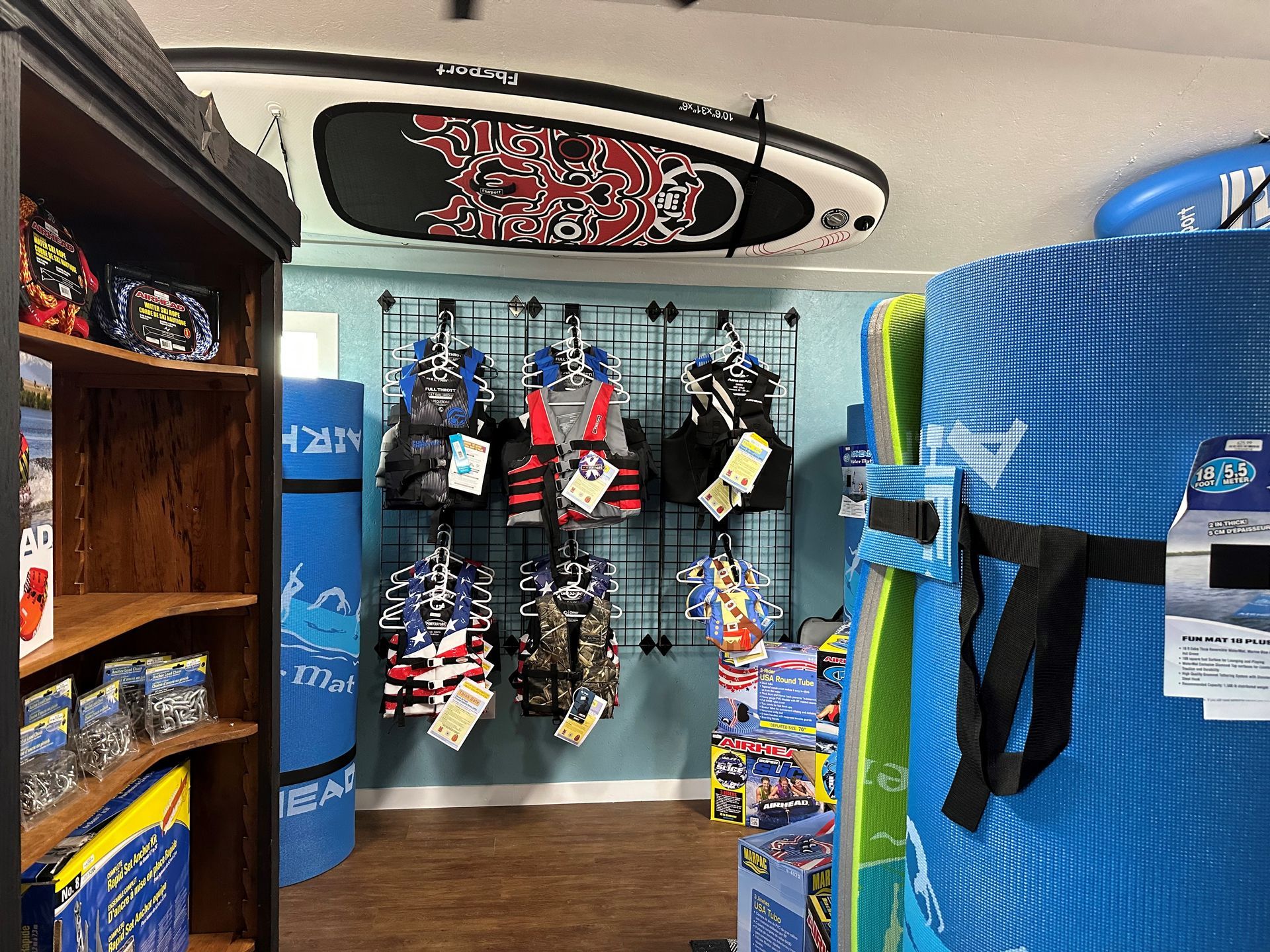 A retail store display featuring life jackets hanging on a grid wall, blue foam water mats, and a paddleboard on the ceiling.