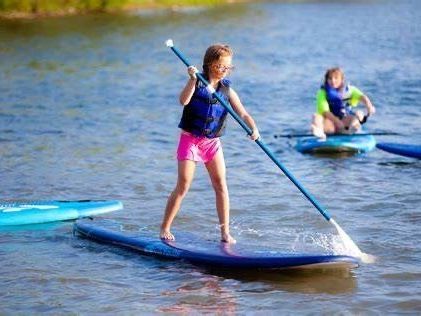 A child in a blue life vest paddleboarding on calm water, with another person paddleboarding in the background.