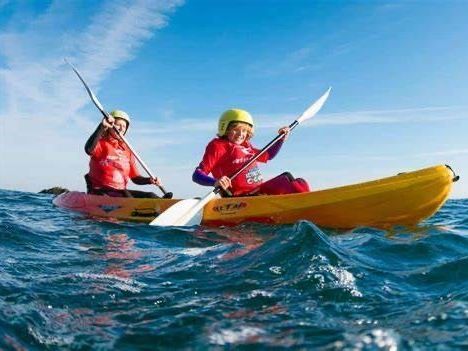 Two people in yellow helmets and red shirts paddling a yellow tandem kayak across choppy blue water under a clear sky.